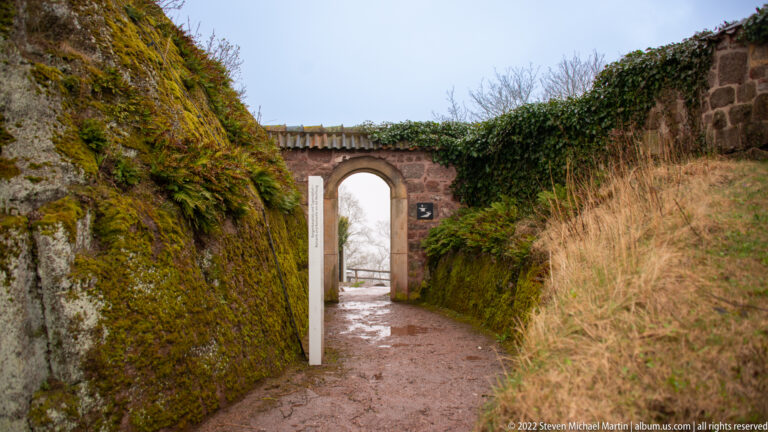 Narrow stone-lined path leading through a small arch, with mossy rock on the left and ivy-covered wall on the right.