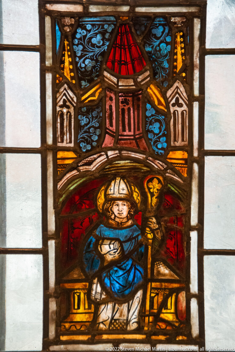 Stained-glass panel of a mitred cleric holding a crozier beneath a colorful architectural canopy.