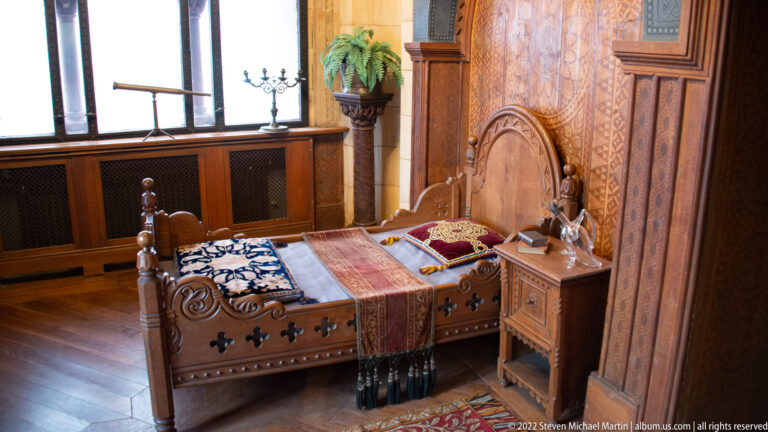 Carved wooden bed with patterned textiles beside a small cabinet in a wood-paneled room with leaded windows.