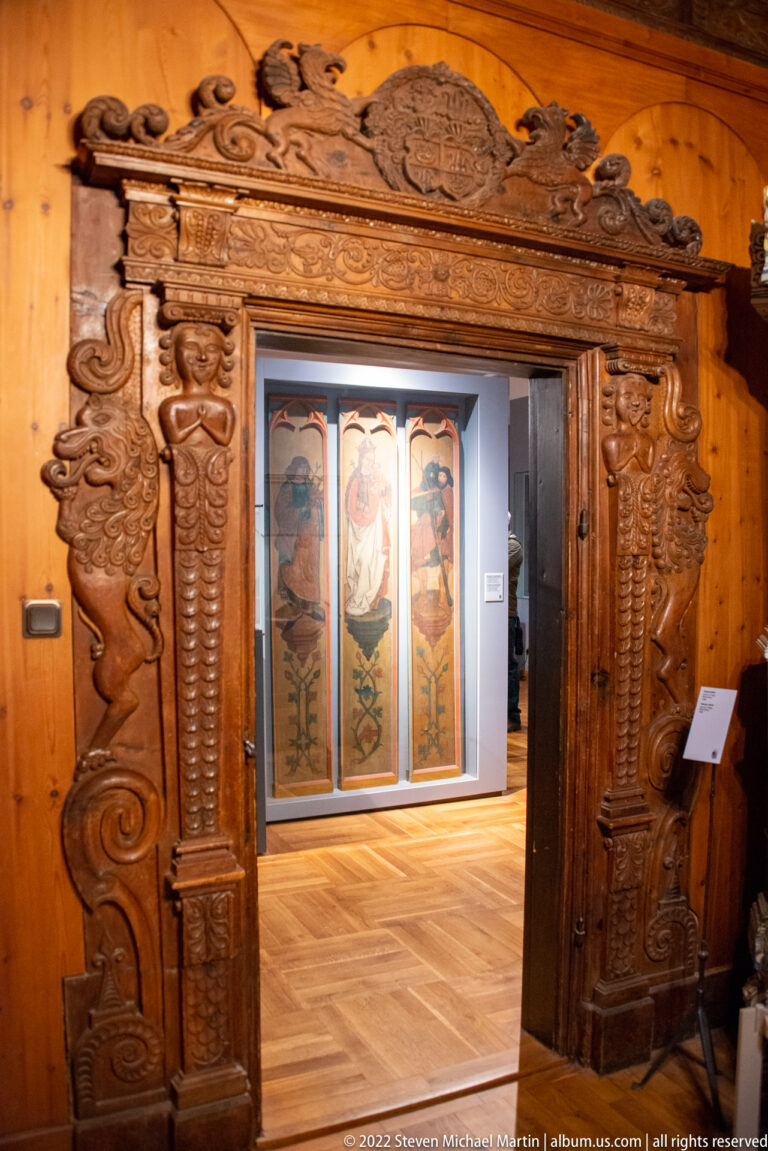Elaborately carved wooden doorway with spiral columns, heraldic crest, sculpted side figures, and painted panels beyond.