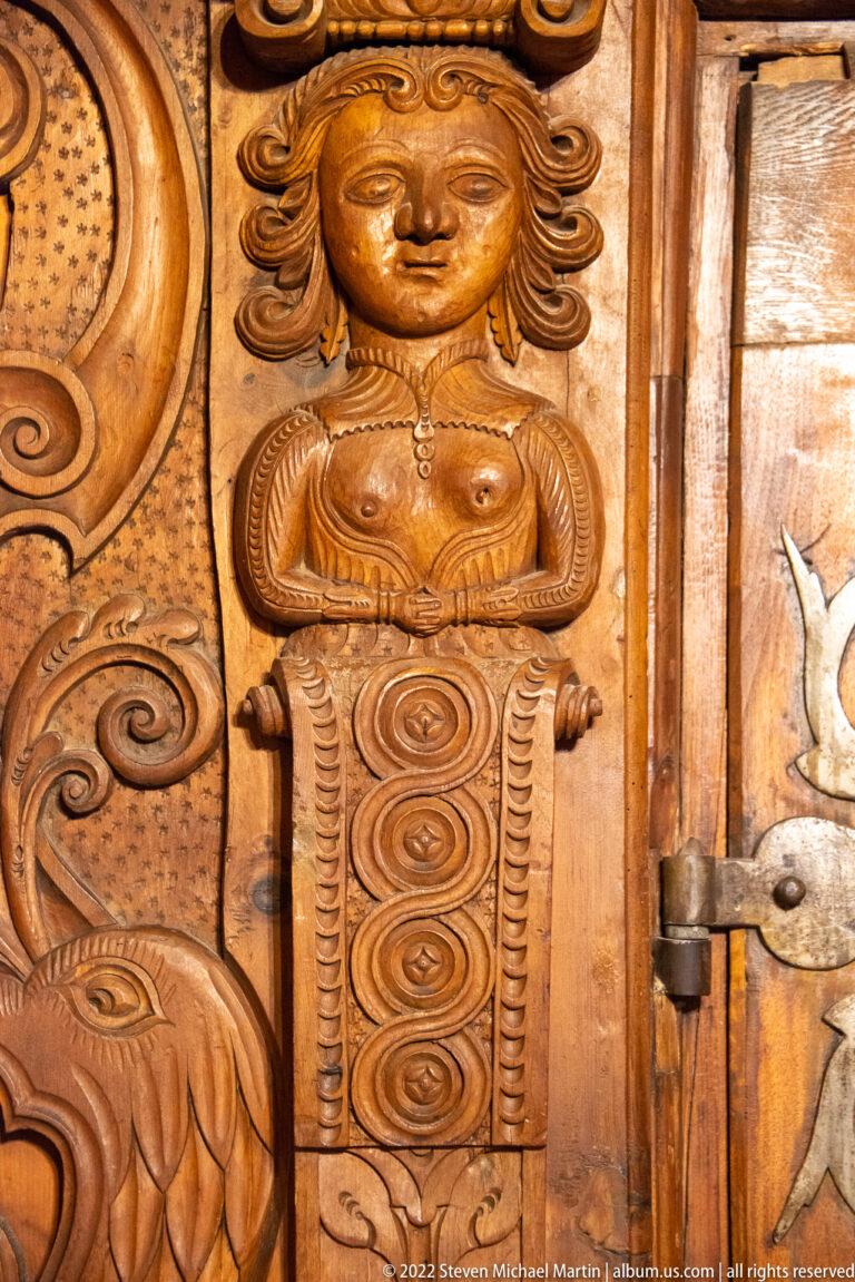 Carved female bust with long hair and folded arms integrated into ornate wooden scrollwork.