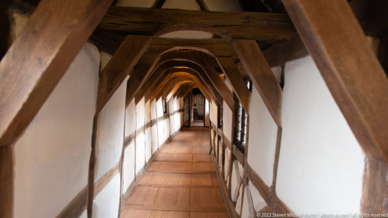 Long narrow gallery with repeated timber braces, white plaster infill, and windows along one side.