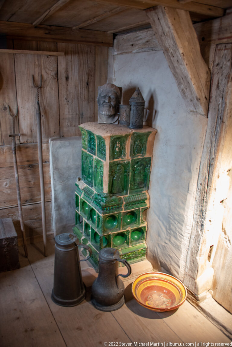 Green glazed-tile stove with dark metal pots and a colorful ceramic dish in a timbered room.