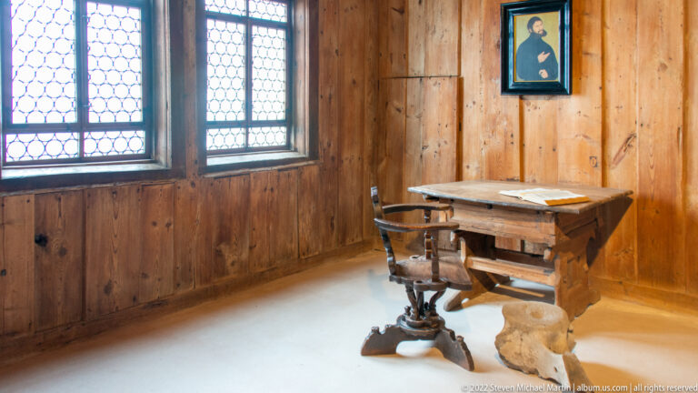 Simple wooden desk and chair beside leaded windows, with a portrait of Martin Luther on the paneled wall.