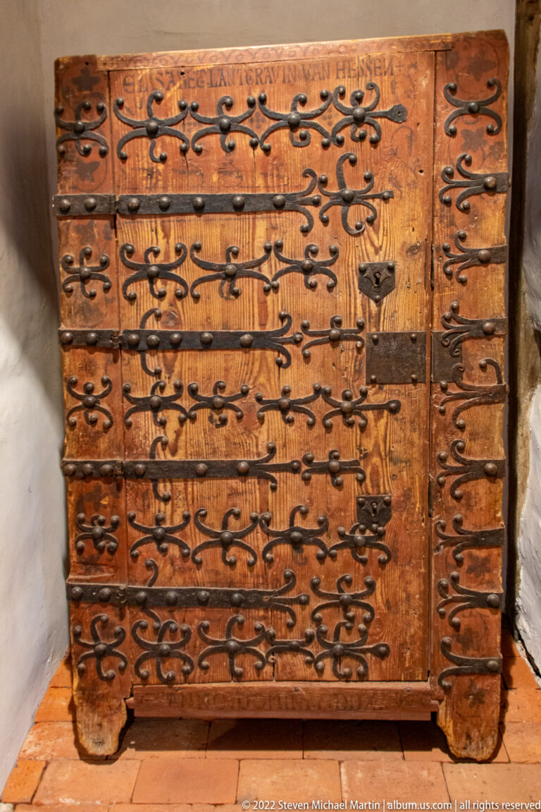 Large wooden chest covered with black iron straps, hinges, scrollwork, and a carved or painted inscription.