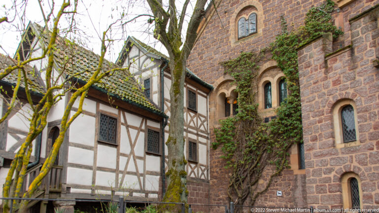 Half-timbered building beside a reddish stone wall with paired round-arched windows and climbing ivy.