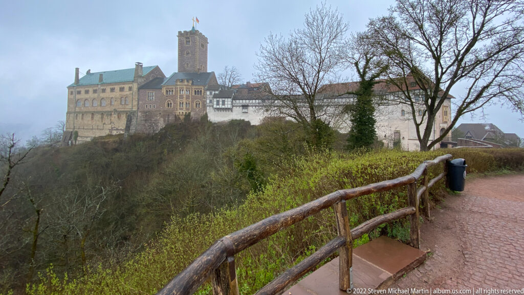 Wartburg Castle on a wooded ridge seen from a curving path with rustic railing in misty weather.
