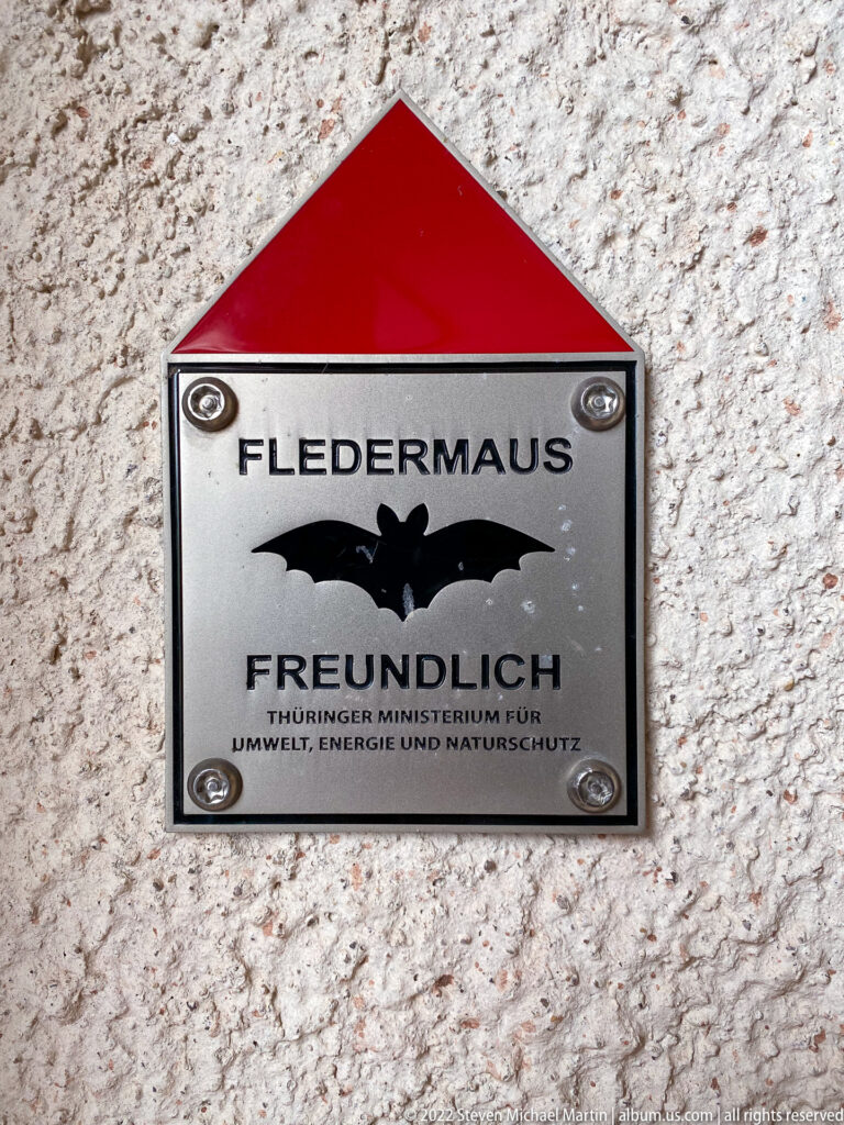 Metal plaque reading 'Fledermaus freundlich' with a black bat silhouette and red roof-shaped top.
