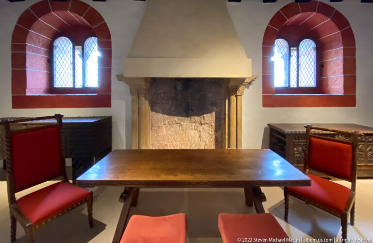 Room with large stone fireplace, wooden table, red-upholstered chairs, and two small leaded windows.