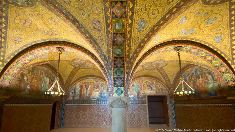 Central stone column beneath intersecting gold mosaic vaults, with narrative scenes and hanging lamps.