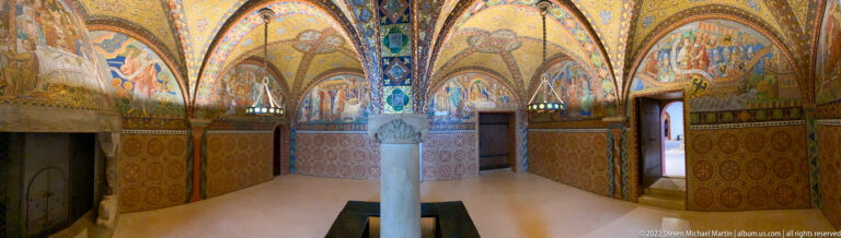 Wide view of the Elisabeth Kemenate with central column, gold mosaics, patterned walls, doorways, and fireplace.