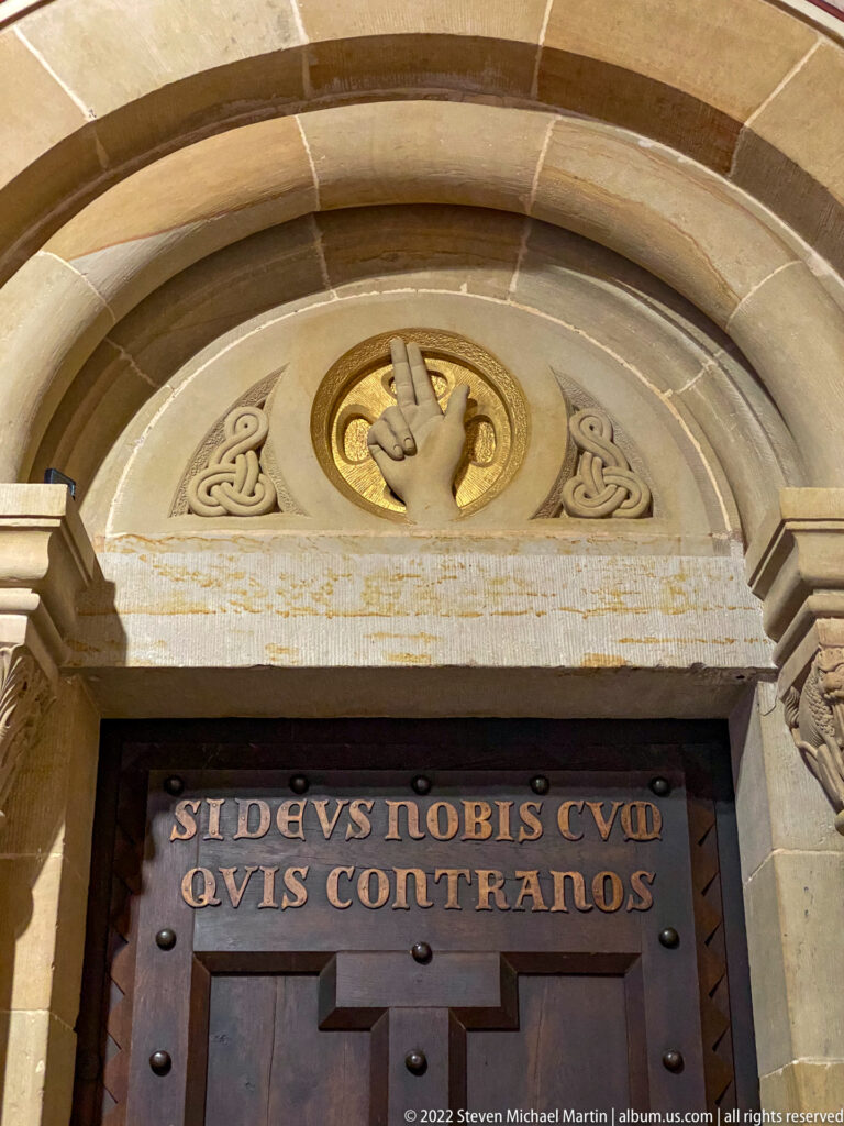 Studded wooden door beneath a round stone arch, inscribed with the Latin phrase 'Si Deus nobis ... quis contra nos.'