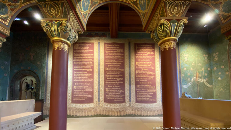 Ornate chamber with painted columns framing three tall inscribed panels against a red wall.