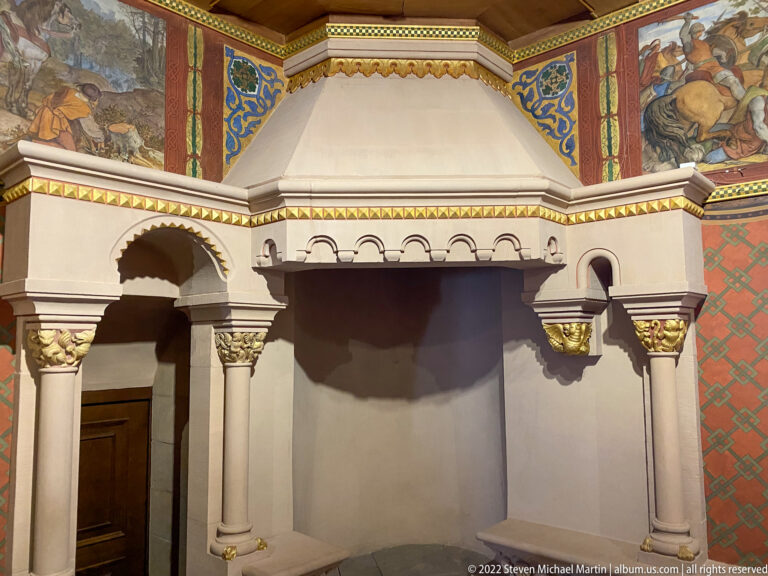 Large hooded fireplace with slender columns, gilded capitals, small arches, and painted decoration above.