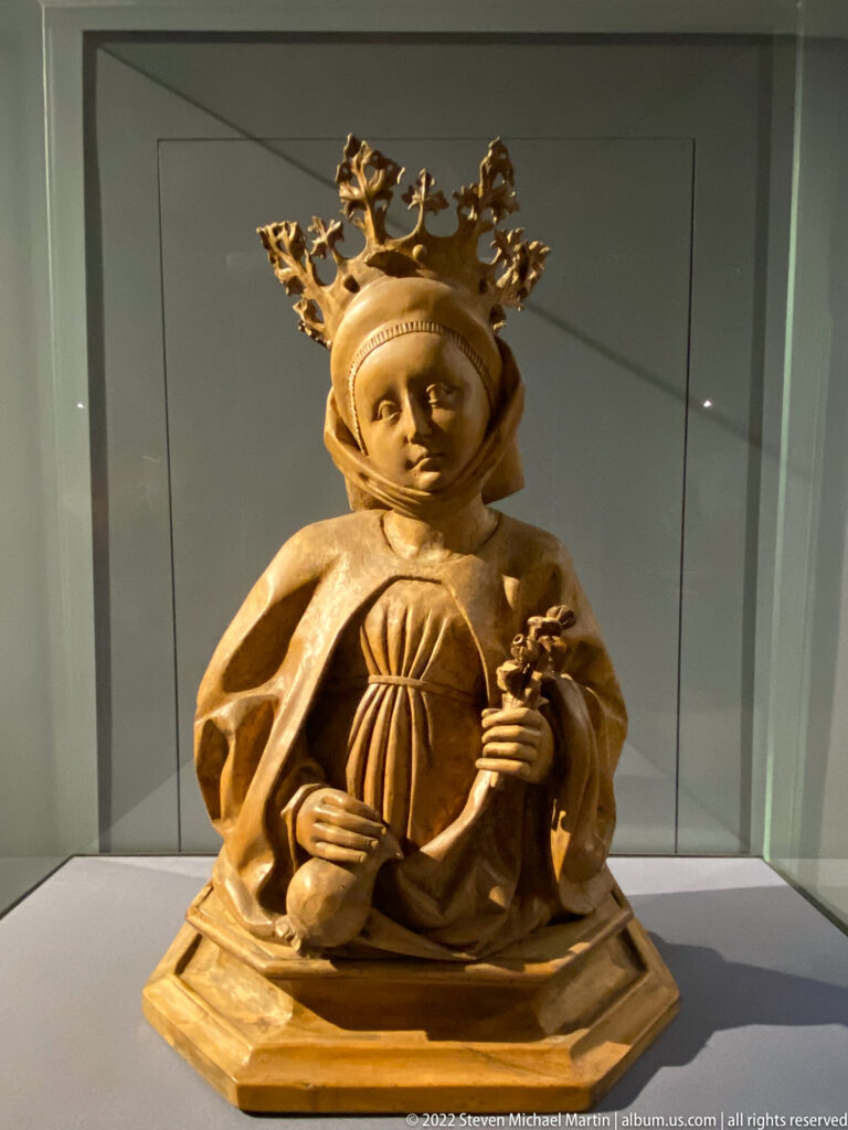 Small carved crowned woman in medieval dress holding a slender object, displayed inside a museum case.
