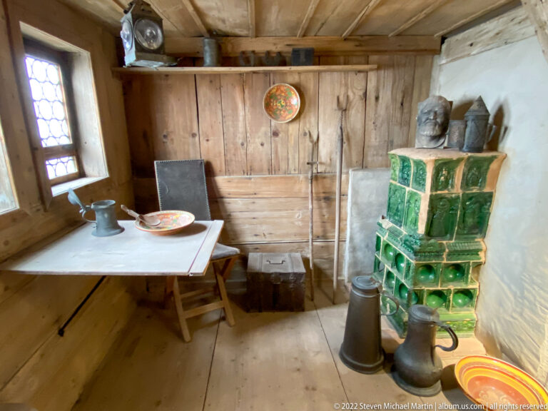 Simple Lutherstube interior with wooden table, chair, green glazed-tile stove, vessels, and leaded window.