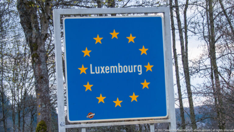 Blue Luxembourg border sign encircled by European Union stars beside a wooded roadside.