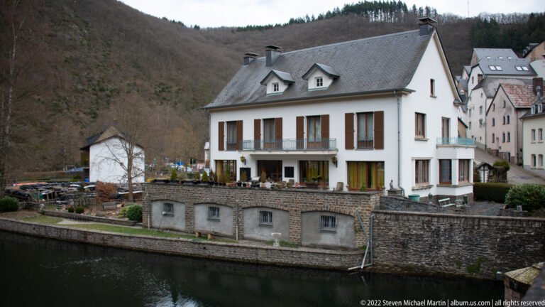 White riverside hotel building with dark shutters, slate roof, dormers, terrace, and stone embankment in Esch-sur-Sûre.