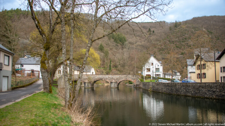 Low multi-arch stone bridge reflected in the Sûre, with trees and village houses beyond in Esch-sur-Sûre.