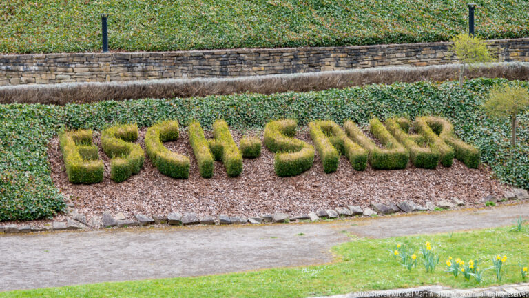 Topiary letters spelling ESCH-SUR-SURE set in reddish mulch and groundcover on a landscaped slope.