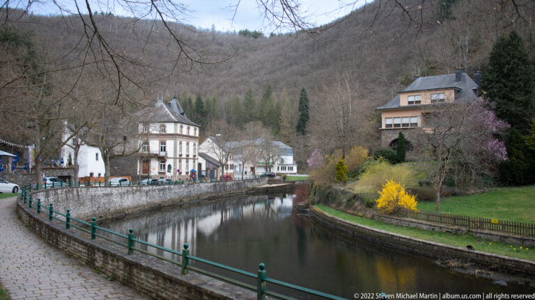 Sûre river channel curving between stone embankments, village houses, gardens, and wooded hills in Esch-sur-Sûre.