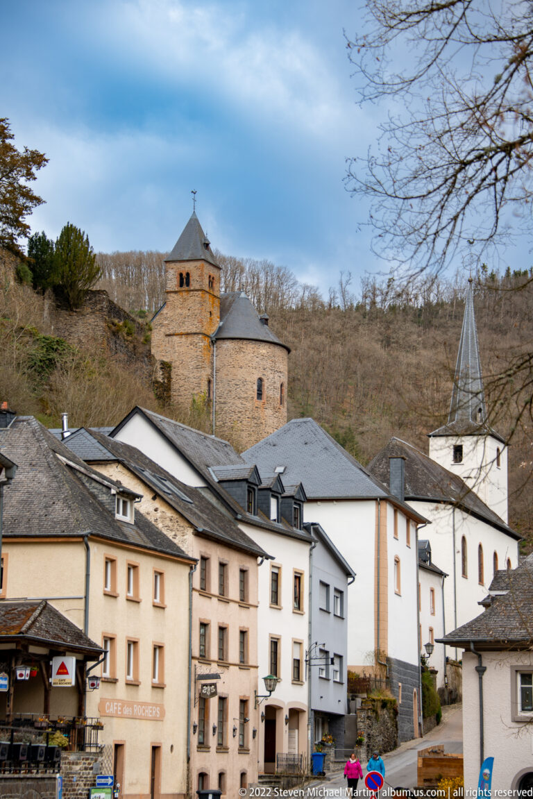 Stone castle chapel and tower above village roofs, with the white-spired parish church at right in Esch-sur-Sûre.