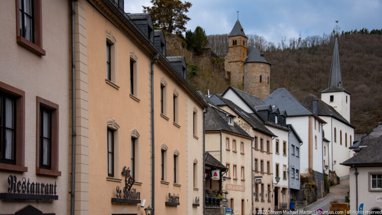 Narrow Esch-sur-Sûre street lined with houses, with the castle chapel and white parish church spire rising behind.