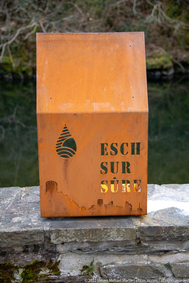 Rust-colored metal block marked ESCH SUR SURE with a cut-out emblem, resting on a stone wall.