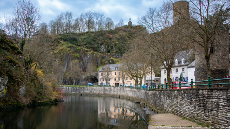 Sûre river beside a stone embankment and cliff, with riverside buildings and castle ruins on the ridge above.
