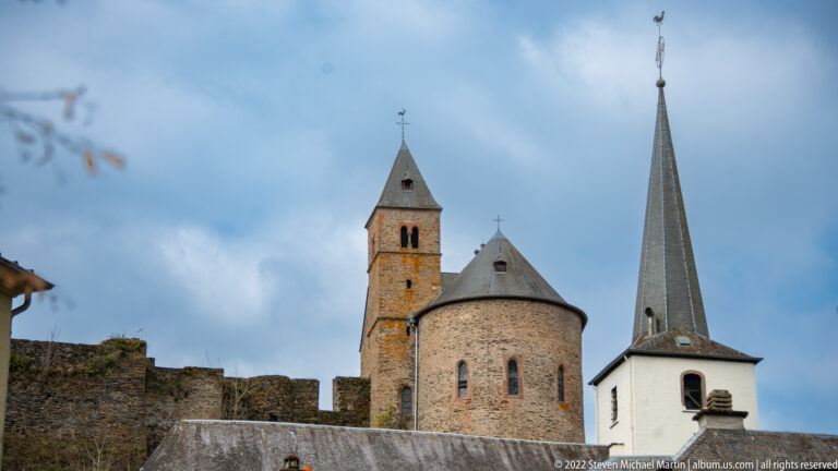 Stone castle chapel with rounded apse beside the white-spired parish church above the castle wall in Esch-sur-Sûre.