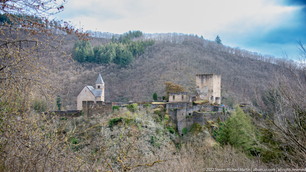 Esch-sur-Sûre Castle ruins on a rocky ridge, with chapel, stone towers, walls, and wooded hills behind.