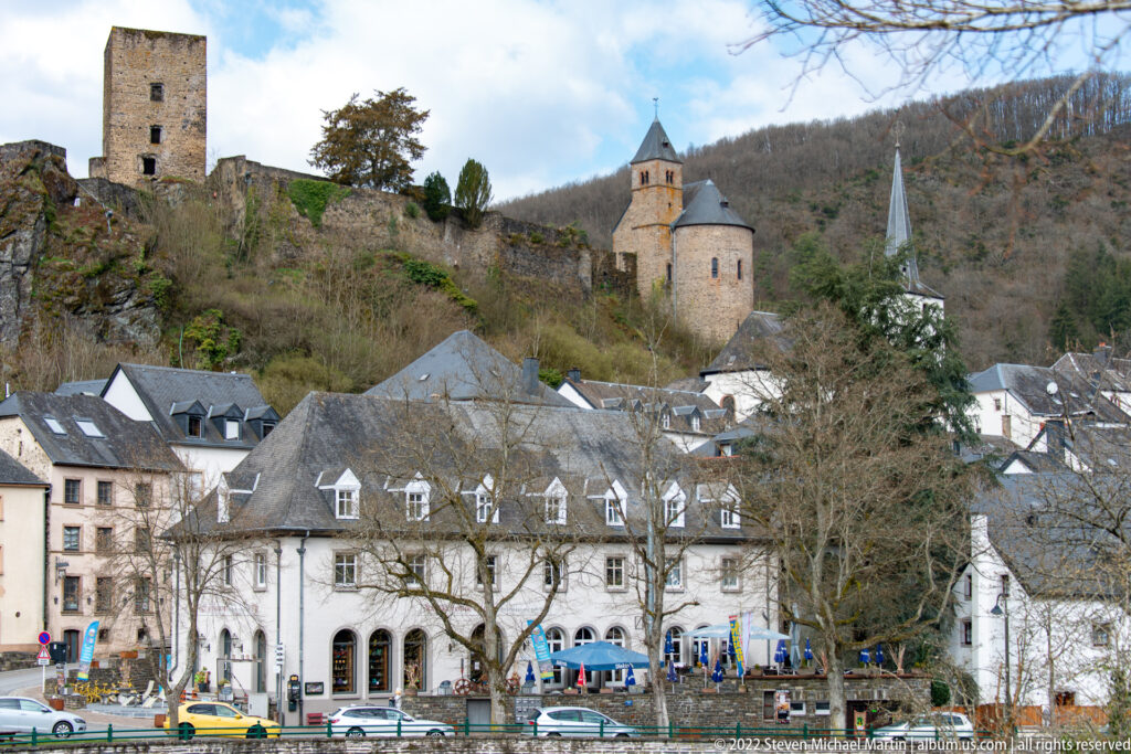 Large pale riverfront building below the Esch-sur-Sûre castle chapel and walls, with parish church spire at right.