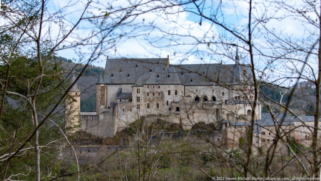 Vianden Castle seen across the valley through bare branches, with steep roofs, gables, towers, and fortified walls.