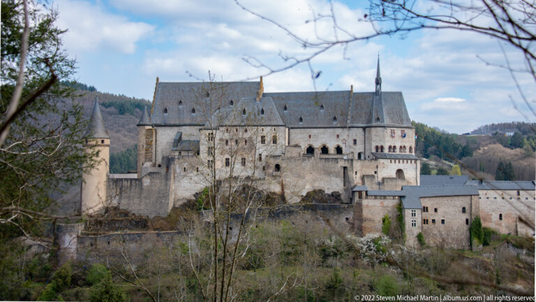 Profile view of Vianden Castle showing high-roofed palace ranges, round towers, lower walls, and buildings descending the slope.