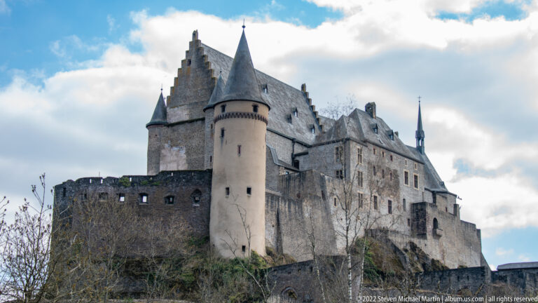 Low-angle view of Vianden Castle with round tower, steep gables, slate roofs, and massive stone walls above the slope.