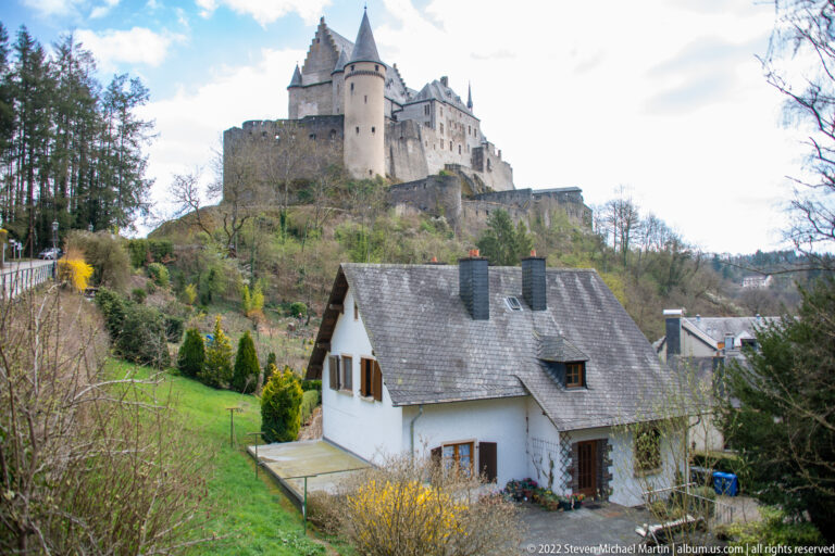 Vianden Castle above a white hillside house and garden, with round tower, high roofs, walls, and trees.