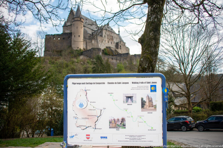 Pilgrimage-route information panel and wayfinding markers in the foreground, with Vianden Castle rising behind trees.