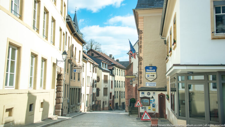 Narrow descending street lined with pale buildings, projecting shop signs, and flags in Vianden's old town.