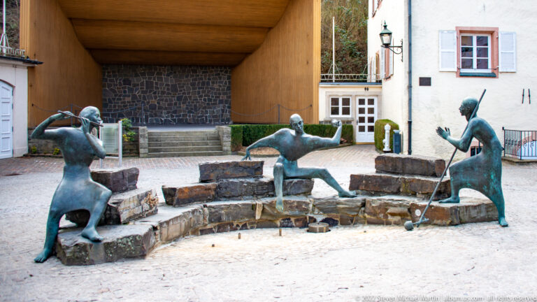 Three weathered bronze human figures stand around a low stone fountain basin in a paved courtyard.