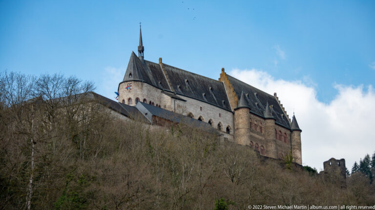 Vianden Castle rises above a leafless wooded hillside, with pale walls, steep roofs, towers, and a slender spire.