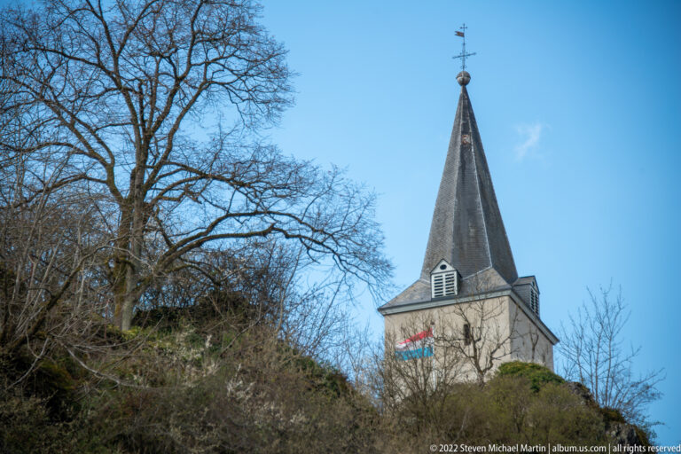 Square pale church tower with louvered belfry and tall dark spire rises beside a steep wooded slope.