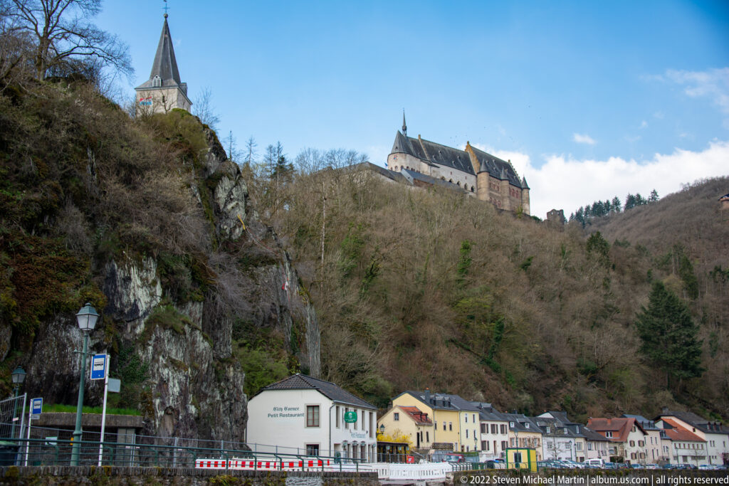 Vianden Castle crowns a wooded rocky hill above small roadside buildings in the valley below.