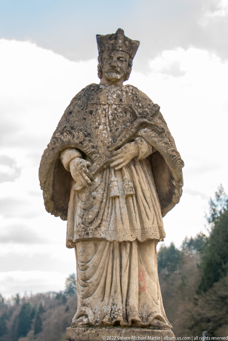Weathered stone statue of a standing figure in a broad mantle and pleated historical dress, hands joined at the waist.