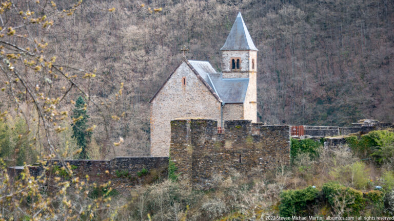 Restored stone chapel at Esch-sur-Sûre Castle behind low ruined walls, with a pointed bell tower and wooded hillside.
