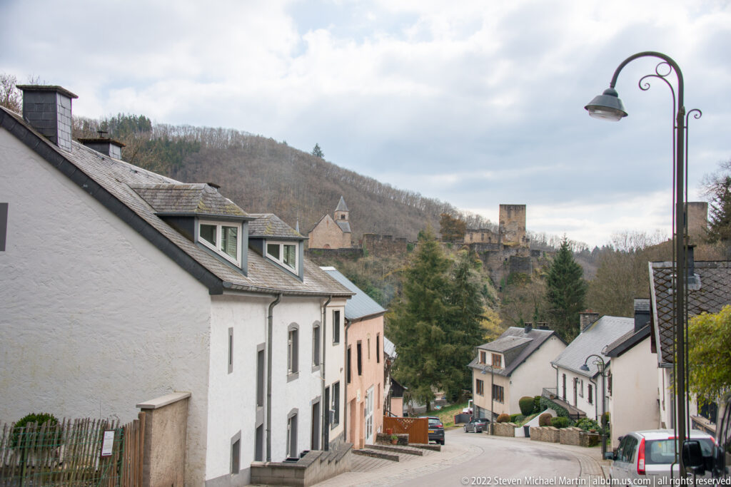 Sloping village street in Esch-sur-Sûre with white houses and the castle chapel and tower visible on the ridge ahead.