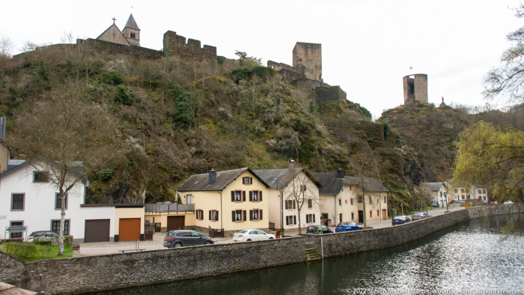 Esch-sur-Sûre Castle ruins above riverside houses and the Sûre, with wooded slopes enclosing the village.
