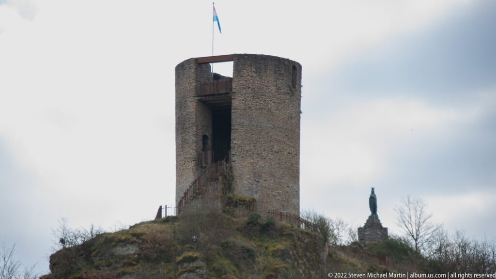 Stone Lochturm watchtower on the Esch-sur-Sûre castle ridge, with Luxembourg flag and a large vertical opening.
