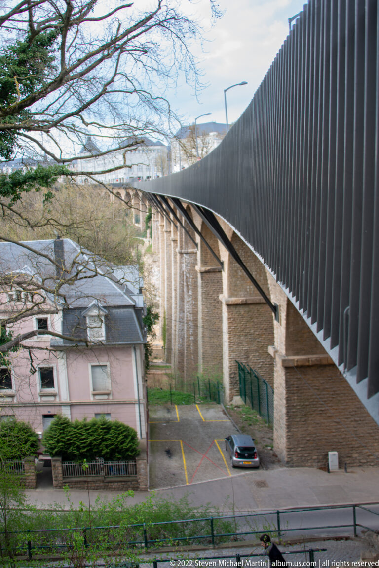 Stone-arch viaduct with a tall dark metal parapet curves above a small building and road at the wooded valley edge.