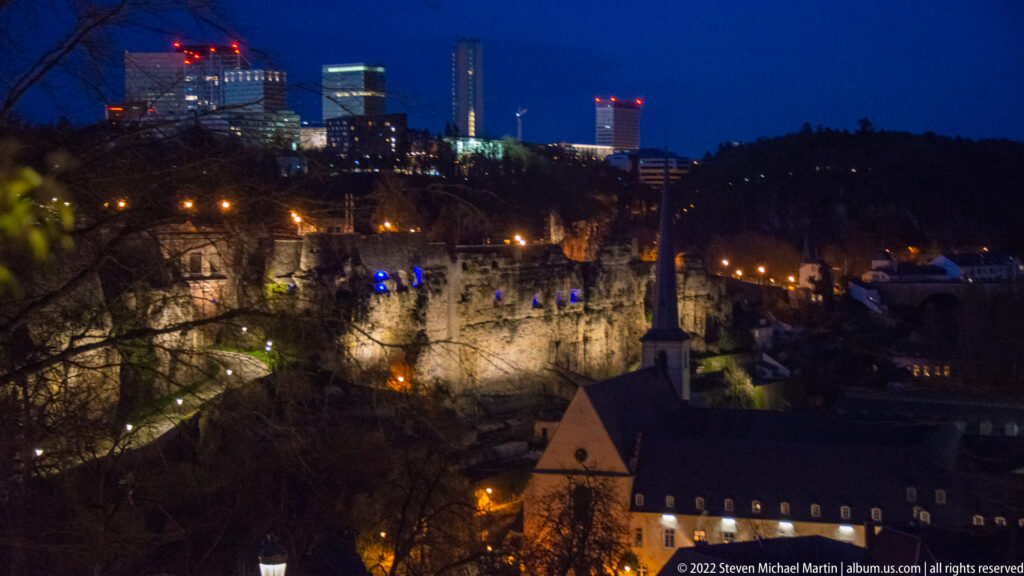 Illuminated fortified cliffs rise above a lower historic building with modern towers glowing on the distant plateau.