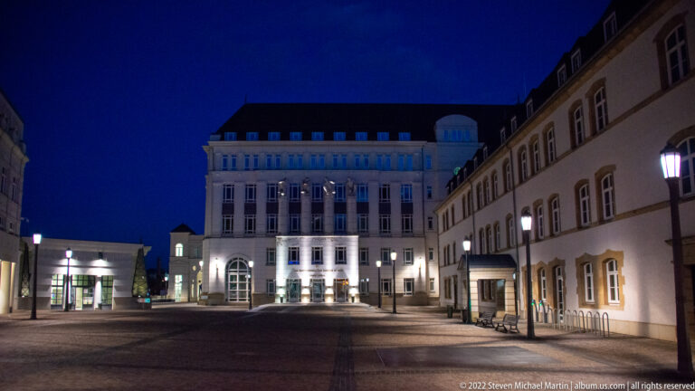 Quiet paved courtyard at night enclosed by pale formal buildings with tall windows and restrained architectural lighting.